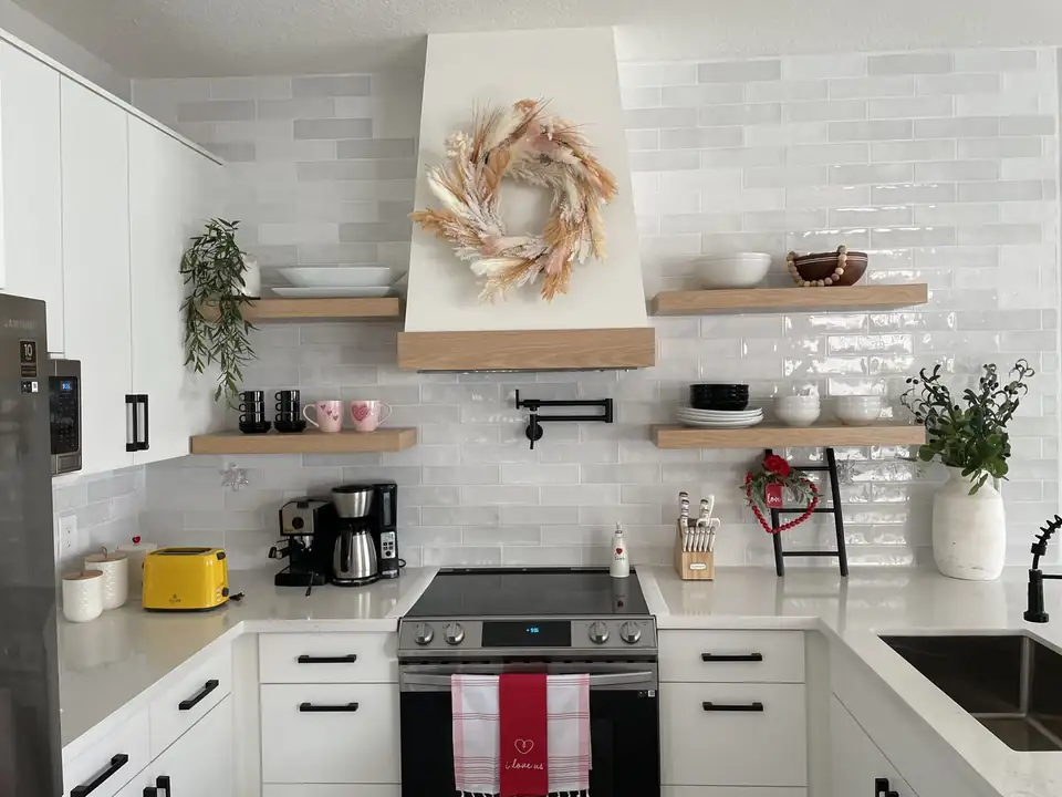Kitchen remodeling by Homestead, FL company Florida Cabinet Solutions: white kitchen with a custom vent hood trimmed in white oak, floating oak shelves, glossy white tile and a pot filler