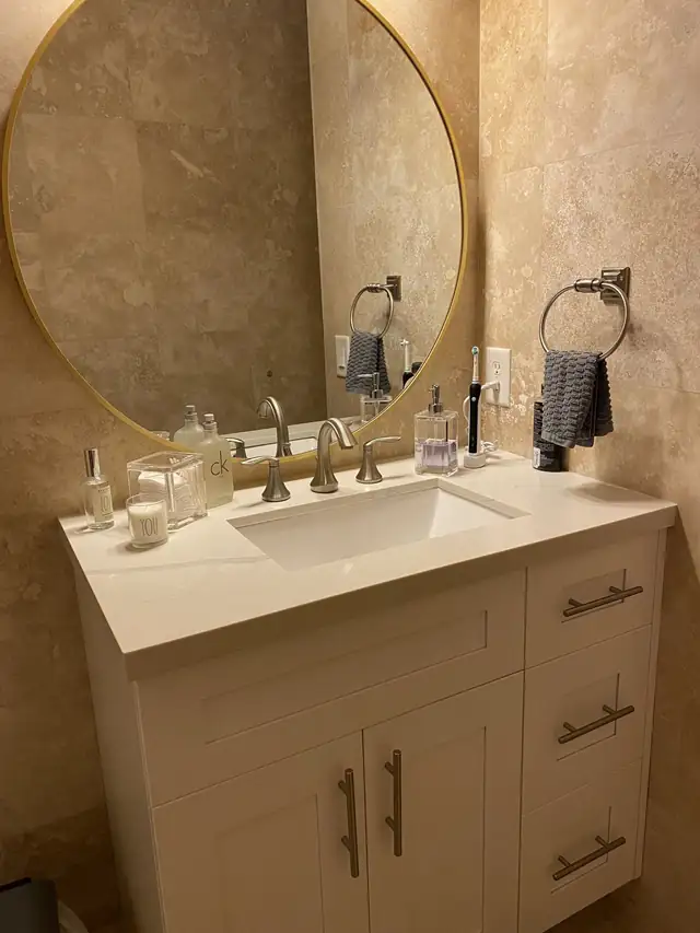 Bathroom vanity with a white countertop, undermount sink and brushed gold faucet below a round mirror